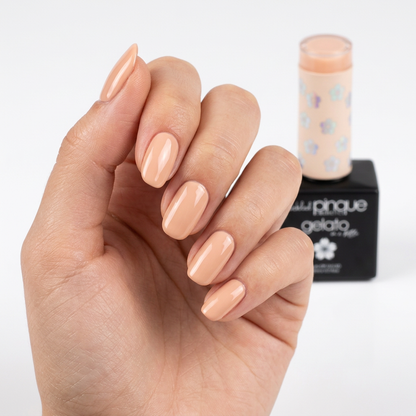 A close-up of a hand with neatly manicured nails painted in a glossy light peach-beige shade, with a tube and black box of nail polish standing behind it on a white background.