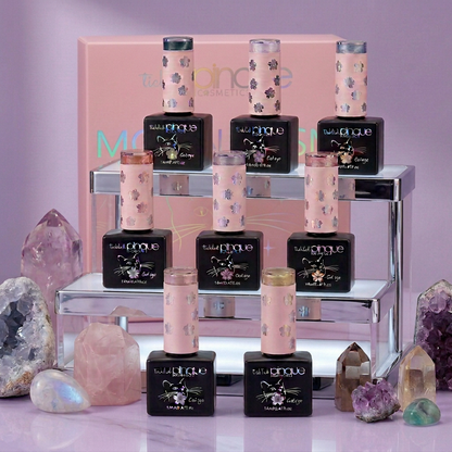 Nine bottles of Tangle Teeze Magic Color Gel Nail Polish in the 'Moonlight Serenity' shade, arranged on a mirrored two-tiered stand with a pink Tangle Teeze box in the background, surrounded by decorative pink and purple crystals on a light purple surface.