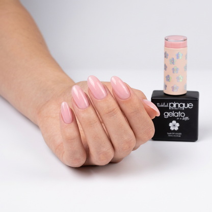 A hand with long, glossy light pink manicured nails is shown next to a bottle of Tasselid Pinque Gelato nail polish on a clean white background.