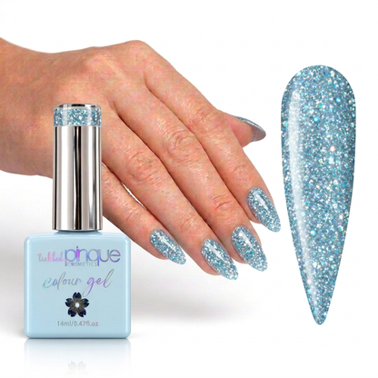 A hand with glittery light blue nail polish is shown beside a bottle of Tula Nova Pastellifica Colour Gel and a large nail swatch displaying the same sparkling blue finish on a white background.